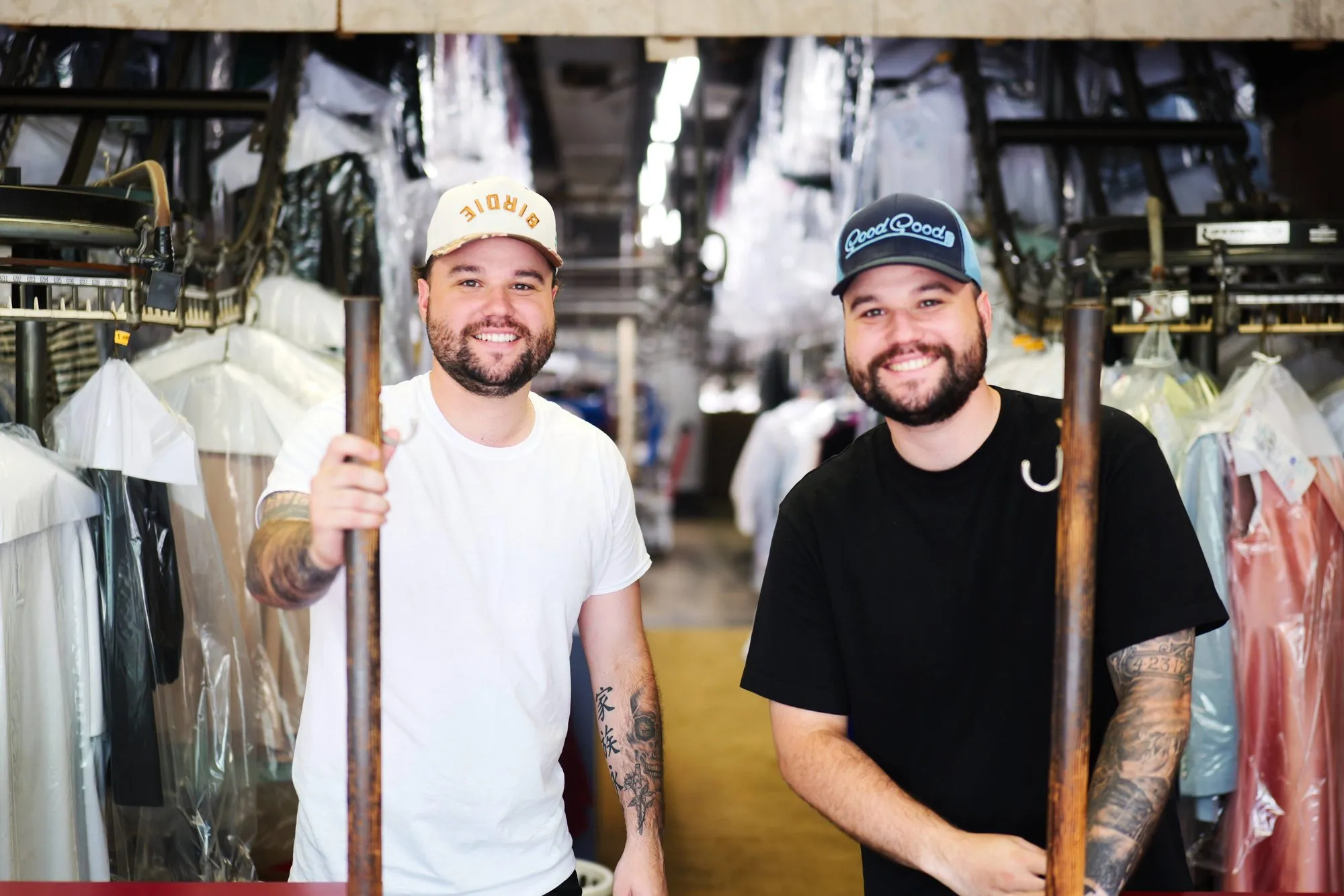 Two of the family standing between long rails of finished, covered garments in the back of the shop.