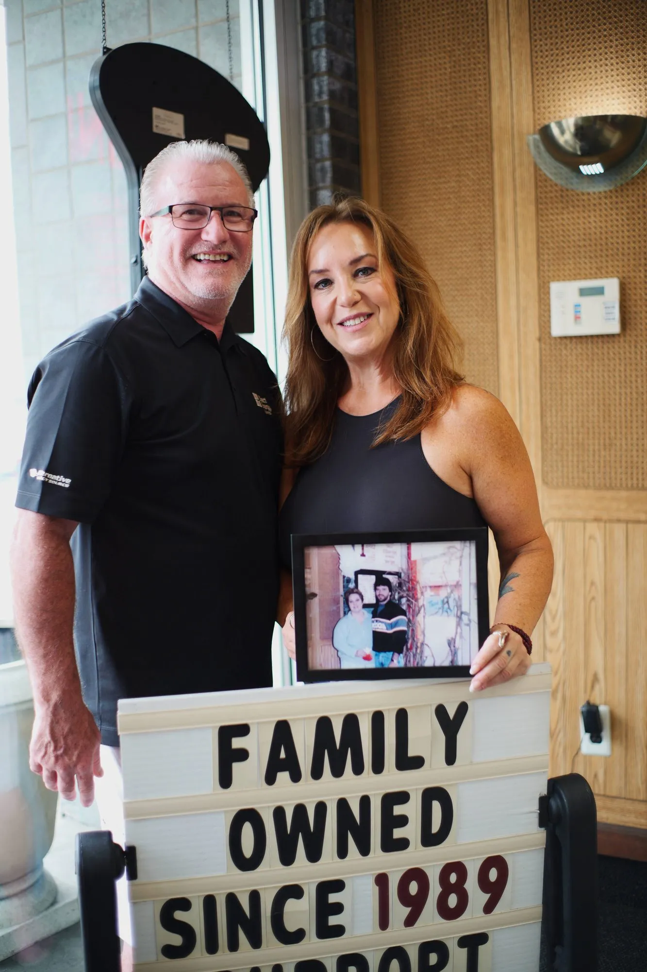 Two of the owners standing in the shop holding a hand-lettered board reading FAMILY OWNED SINCE 1989.