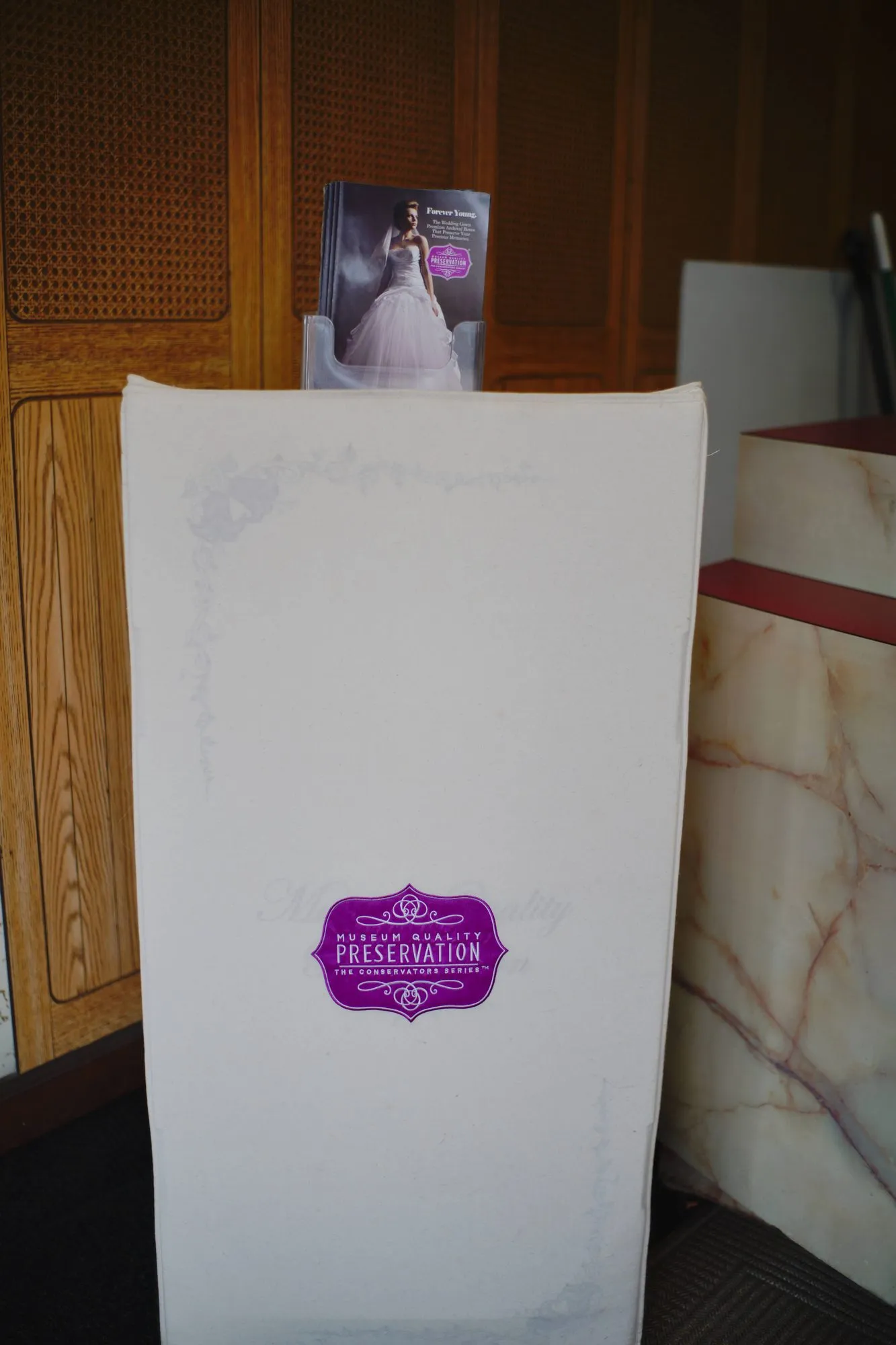 A tall wedding gown preservation box standing at the counter, labelled Museum Quality Preservation, The Conservators Series.