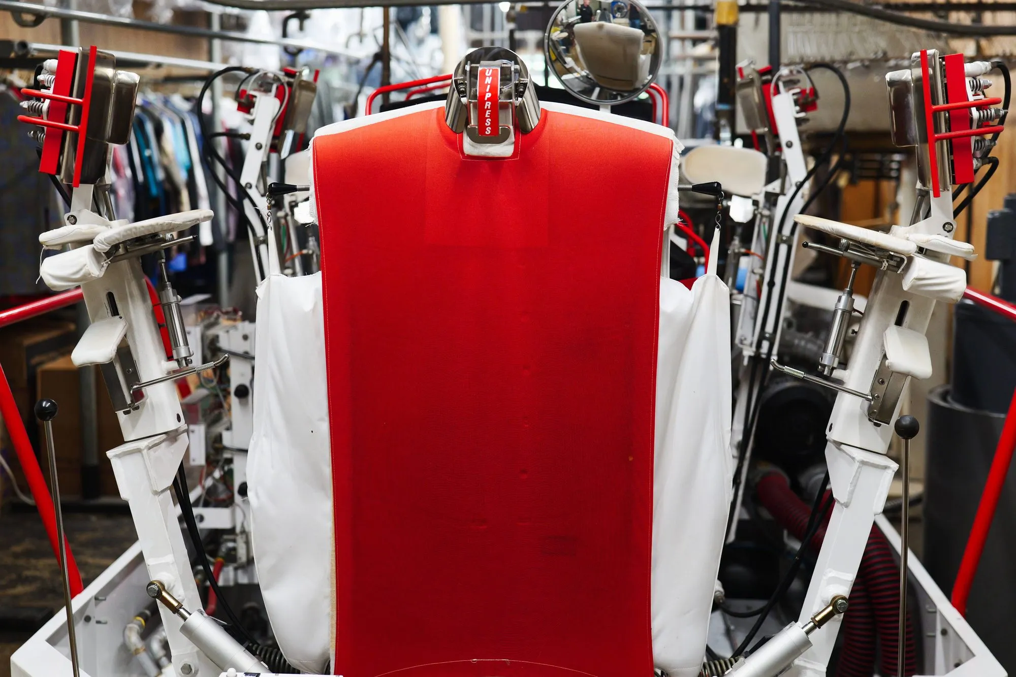 A red-topped shirt press standing among rails of finished garments in plastic covers.