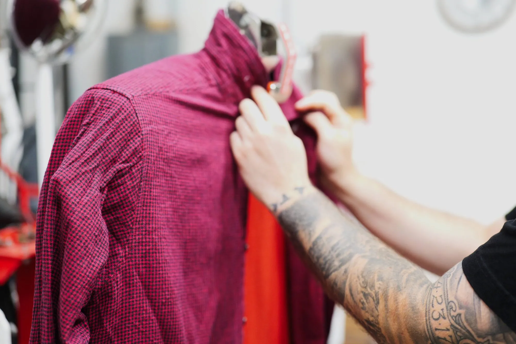 A presser's hands fastening the collar button of a dark red checked shirt on the finishing form.