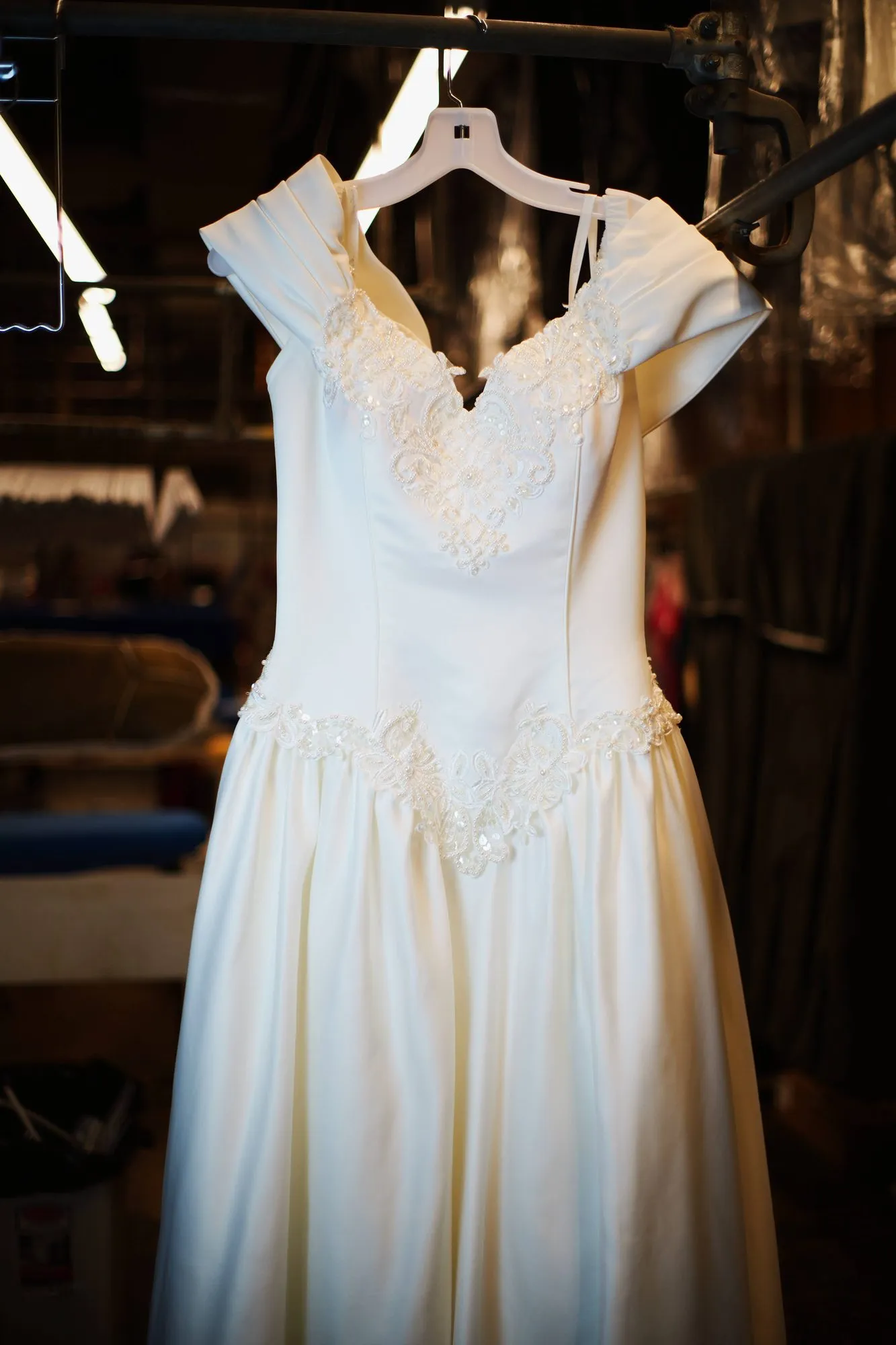 An ivory satin wedding gown with beaded lace at the neckline, hanging on a padded hanger on a rail in the shop.